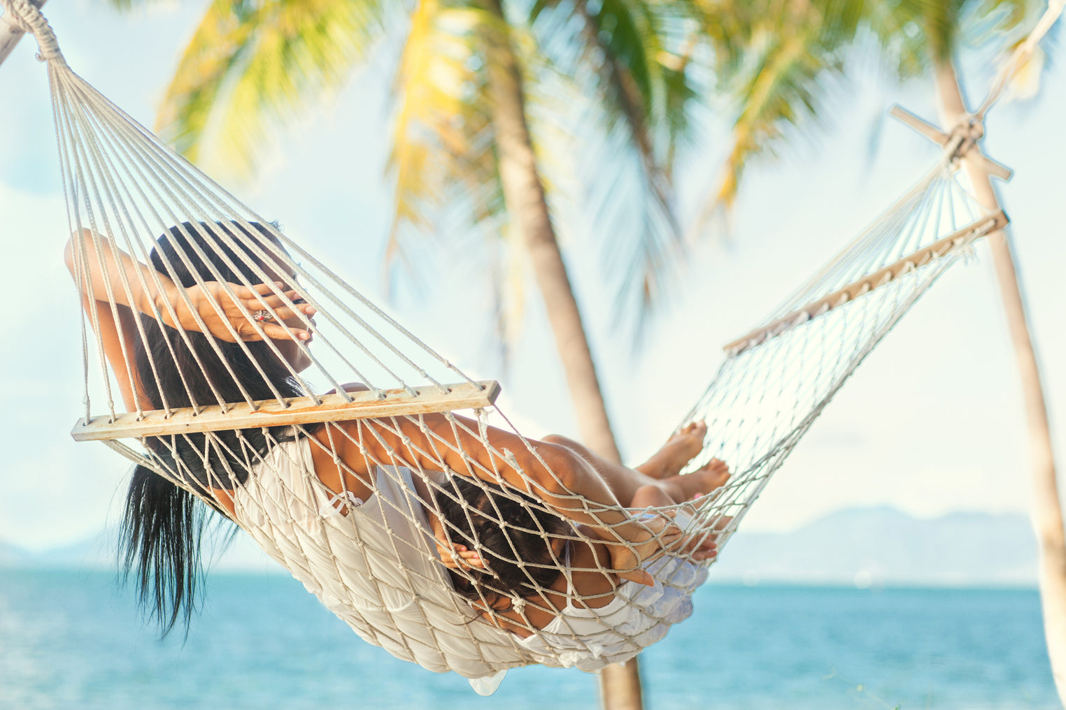 Woman with long black hair lounging on a rope hammock while looking at a tropical beach view.